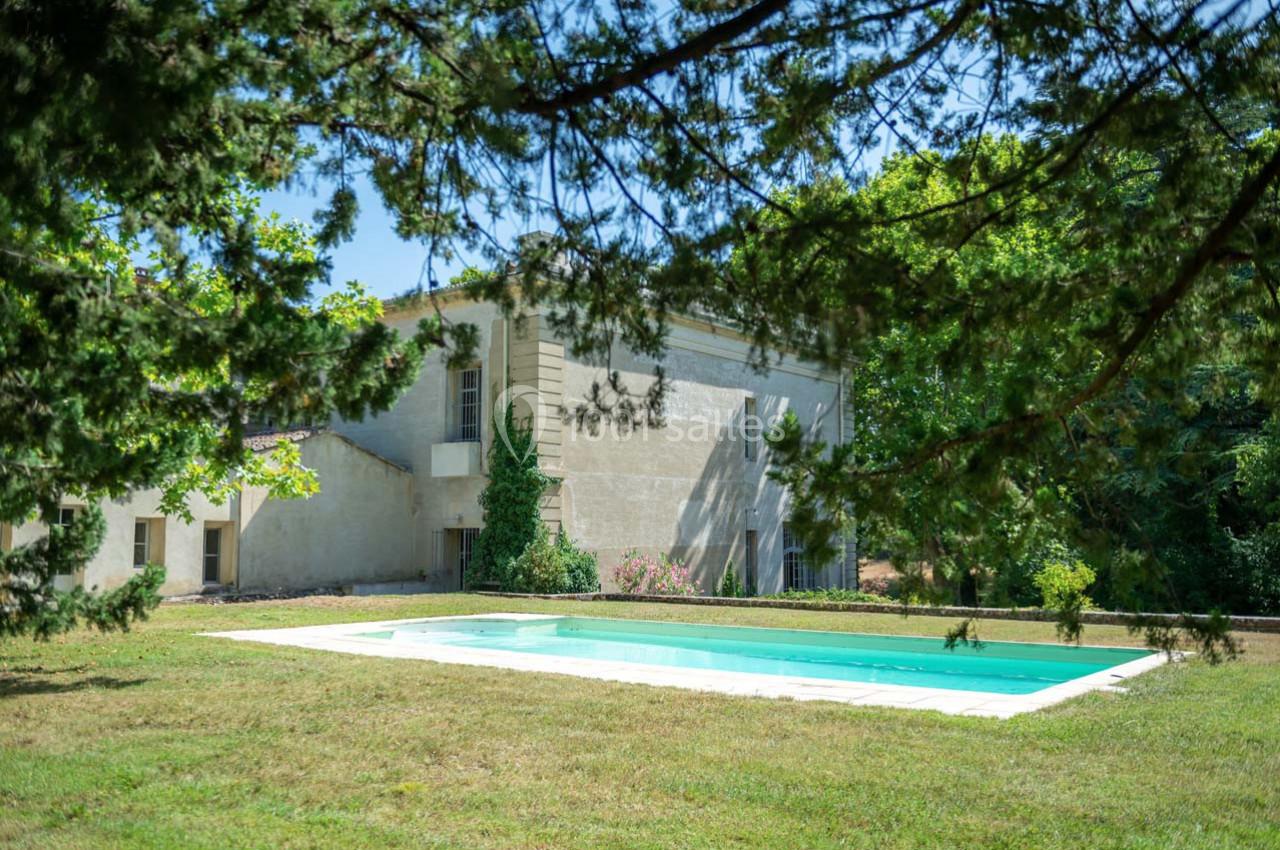 Piscine extérieure entourée de pelouse et d'arbres, située près d'un bâtiment en pierre sous un ciel dégagé.