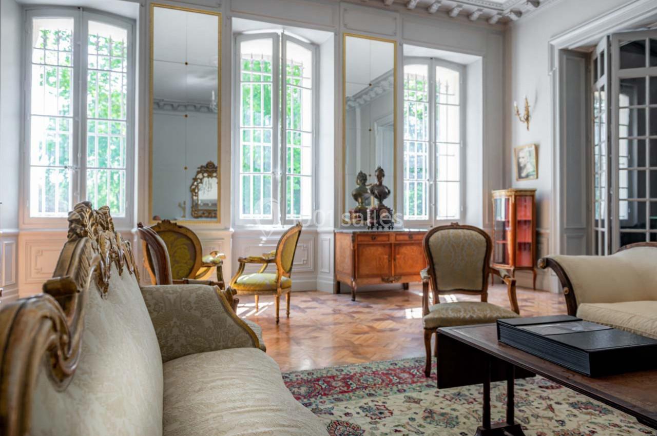 Salon élégant avec mobilier ancien, grandes fenêtres laissant entrer la lumière, parquet et décorations classiques.