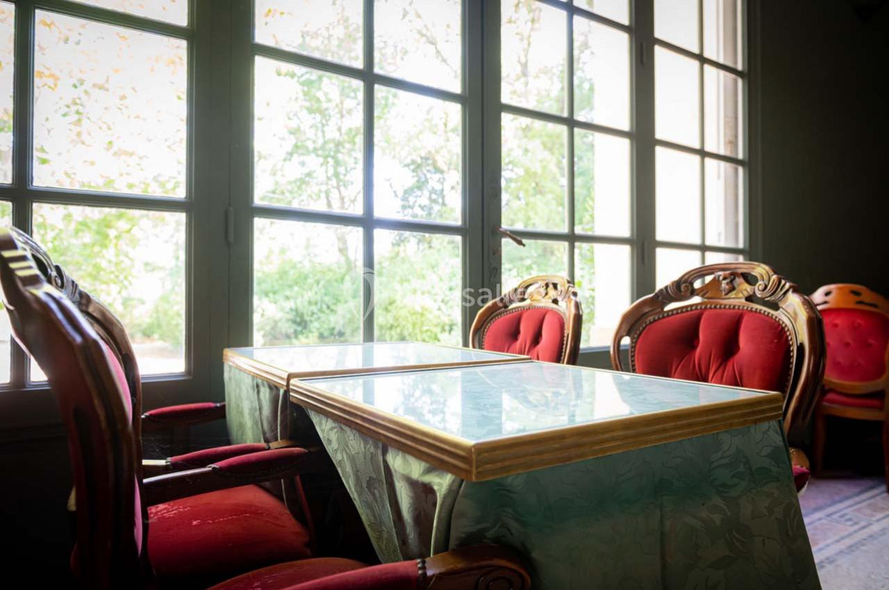 Deux tables recouvertes de nappes vertes entourées de fauteuils rouges, près de grandes fenêtres donnant sur un jardin.
