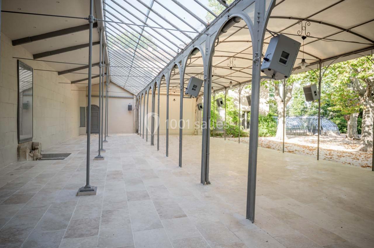 Salle lumineuse avec sol en pierre, structure métallique et toit en verre, ouverte sur un jardin arboré.