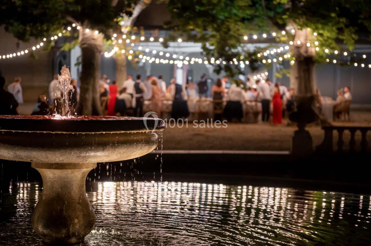 Fontaine éclairée au premier plan avec des invités rassemblés sous des guirlandes lumineuses dans un jardin en soirée.