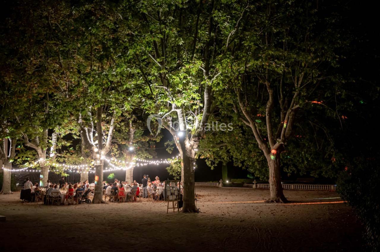 Des convives dînent en plein air sous des arbres illuminés par des guirlandes lumineuses dans un cadre nocturne.