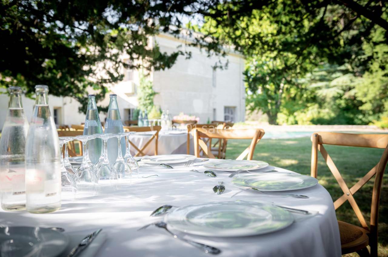 Table dressée en extérieur avec assiettes, couverts, verres et bouteilles, entourée de verdure et d'un bâtiment en arrière…