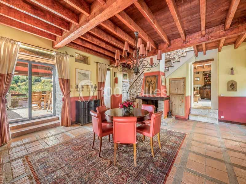 Salle à manger rustique avec poutres apparentes, table ronde entourée de chaises rouges et vue sur une terrasse extérieure.