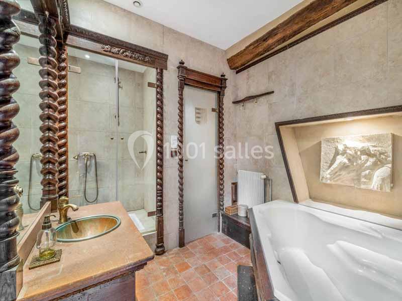 Salle de bain avec baignoire d'angle, douche vitrée, lavabo sur meuble en bois sculpté et murs en carrelage beige.