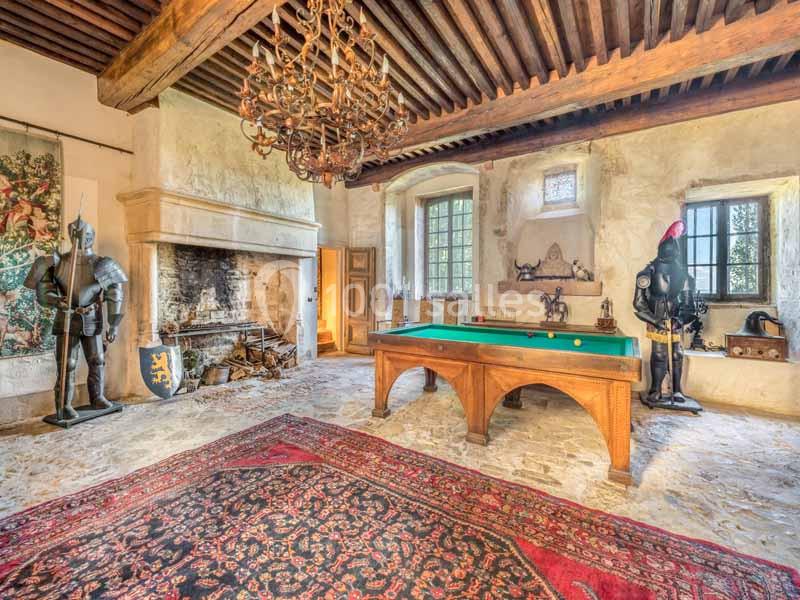 Salle médiévale avec cheminée en pierre, billard, armures décoratives et tapis ancien sur un sol en pierre.