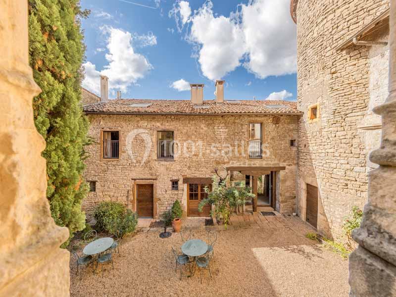Cour intérieure d'une bâtisse en pierre avec tables, chaises, végétation et ciel bleu parsemé de nuages.