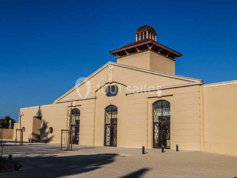 Bâtiment beige avec une tour centrale et des portes vitrées, situé dans une cour dégagée sous un ciel bleu.