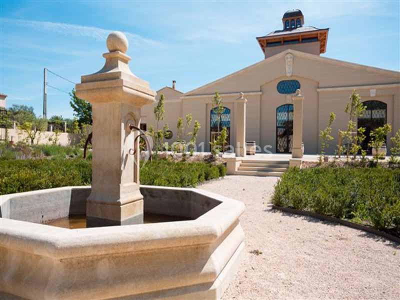 Fontaine en pierre au premier plan devant un bâtiment beige avec des fenêtres cintrées et un toit surmonté d'une tour.
