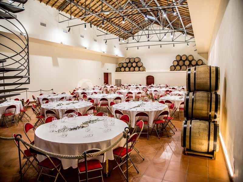 Salle de réception avec tables rondes dressées, chaises rouges et tonneaux en bois empilés contre un mur.