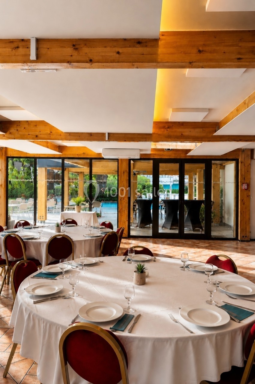 Salle de réception lumineuse avec tables rondes dressées, nappes blanches et vue sur une terrasse extérieure.