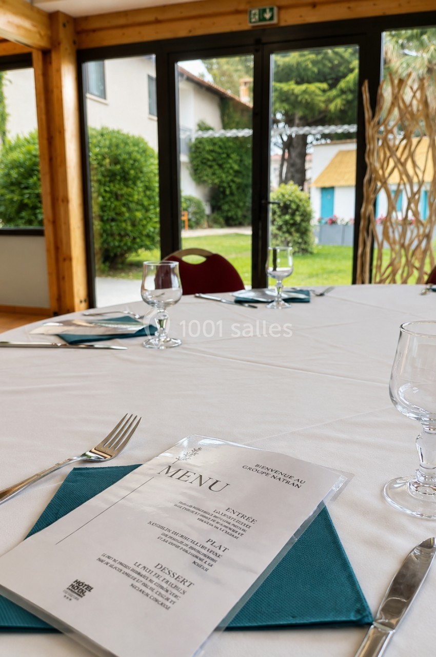 Table dressée avec nappe blanche, menu, couverts et verres, donnant sur un jardin visible à travers de grandes baies vitrées.