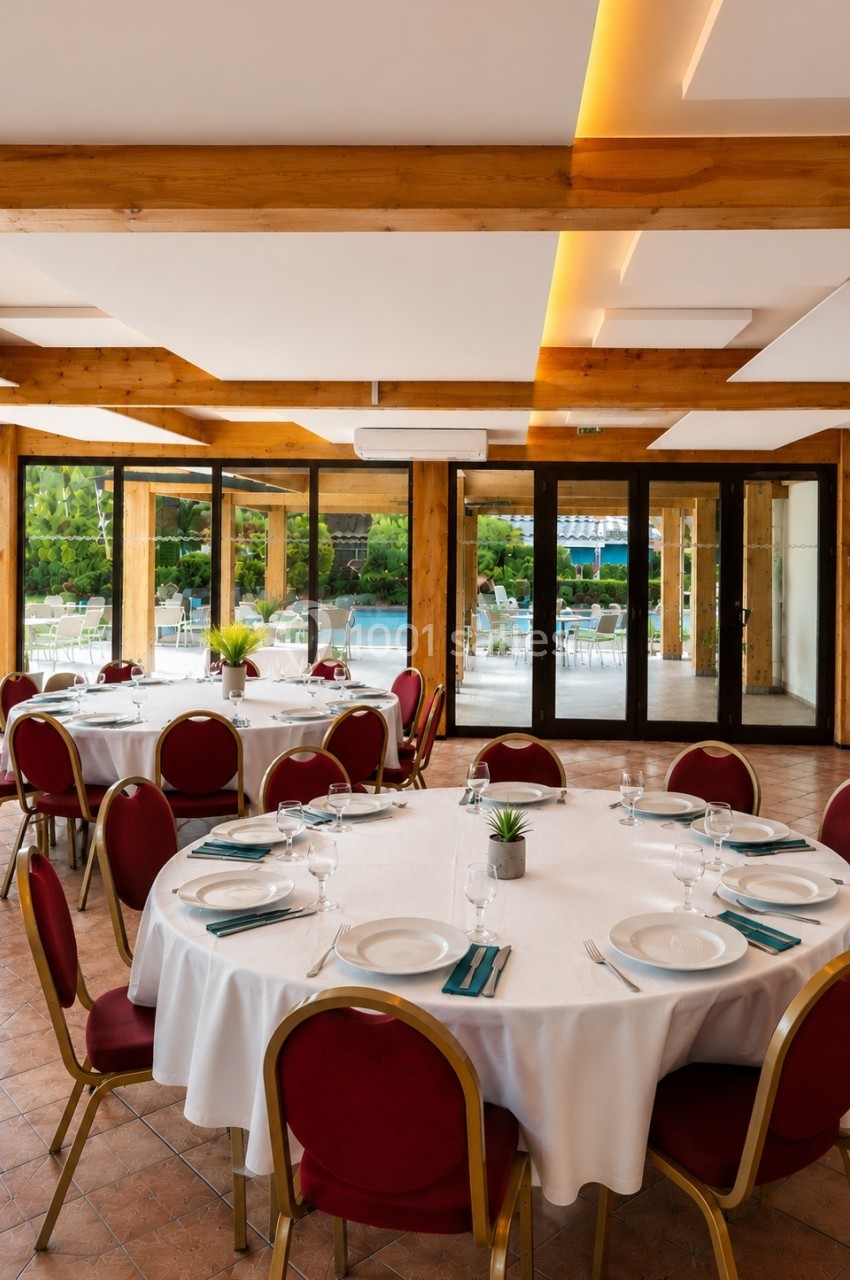 Salle de réception lumineuse avec tables rondes dressées, chaises rouges et vue sur un espace extérieur verdoyant.