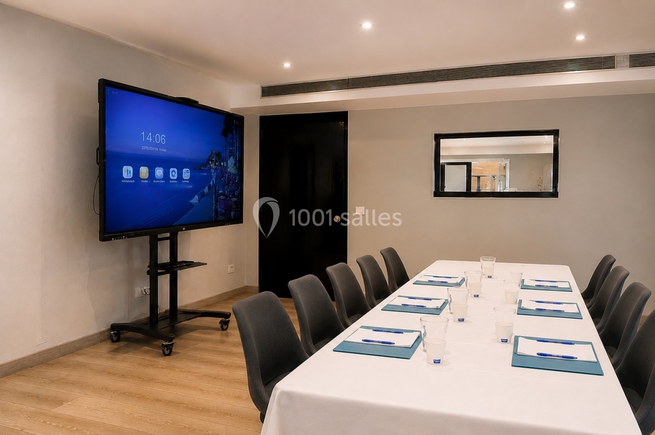 Salle de réunion avec une table rectangulaire dressée, écran interactif allumé et éclairage moderne.
