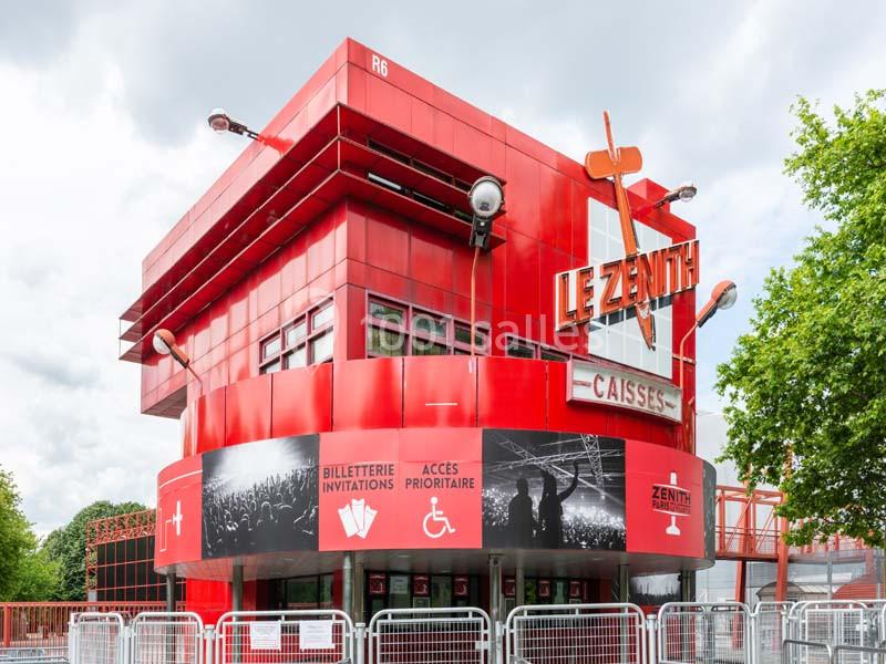 Façade rouge du bâtiment ’Le Zénith’ avec guichets, panneaux d'accès prioritaire et barrières devant l'entrée.