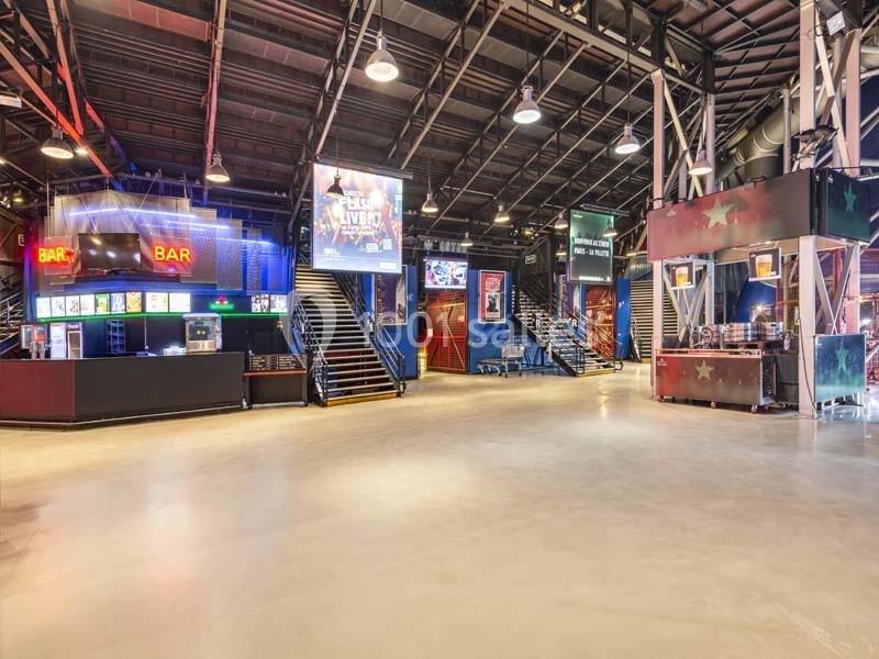 Intérieur d'un espace de loisirs avec bars, écrans lumineux, escaliers et éclairage industriel.