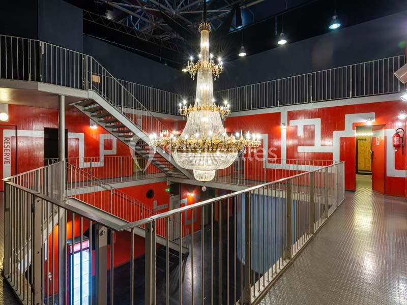 Escalier métallique entourant un grand lustre en cristal dans un espace intérieur aux murs rouges et blancs.