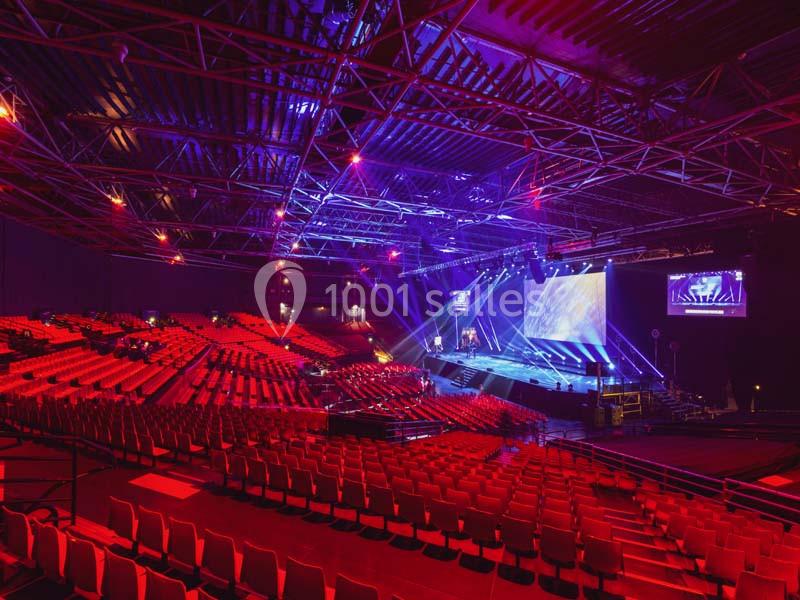 Salle de spectacle vide avec sièges rouges, scène éclairée par des projecteurs et écrans en arrière-plan.