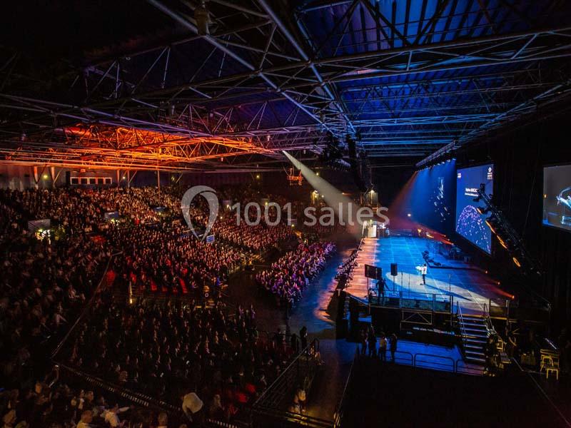 Salle de conférence bondée avec une scène éclairée, un intervenant et un écran géant affichant des visuels.