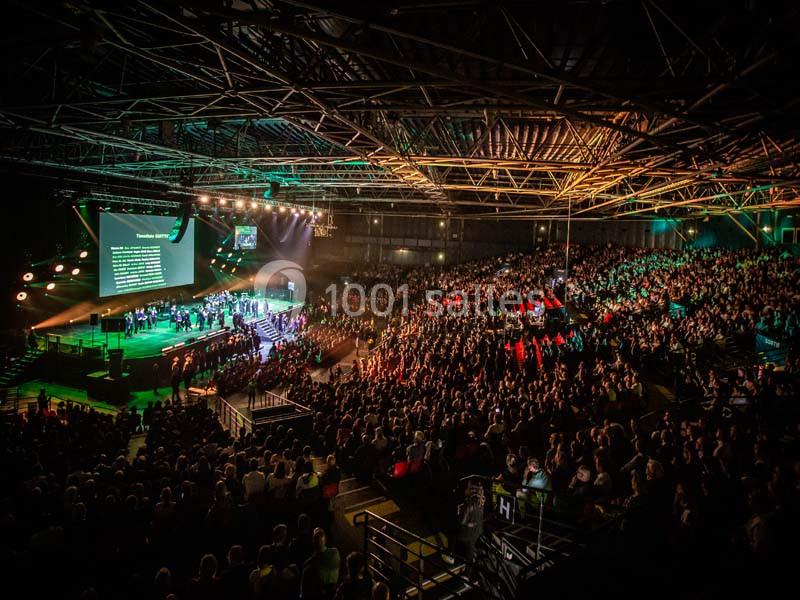 Salle comble lors d'un événement avec scène éclairée, musiciens, écran projetant du texte et public assis.