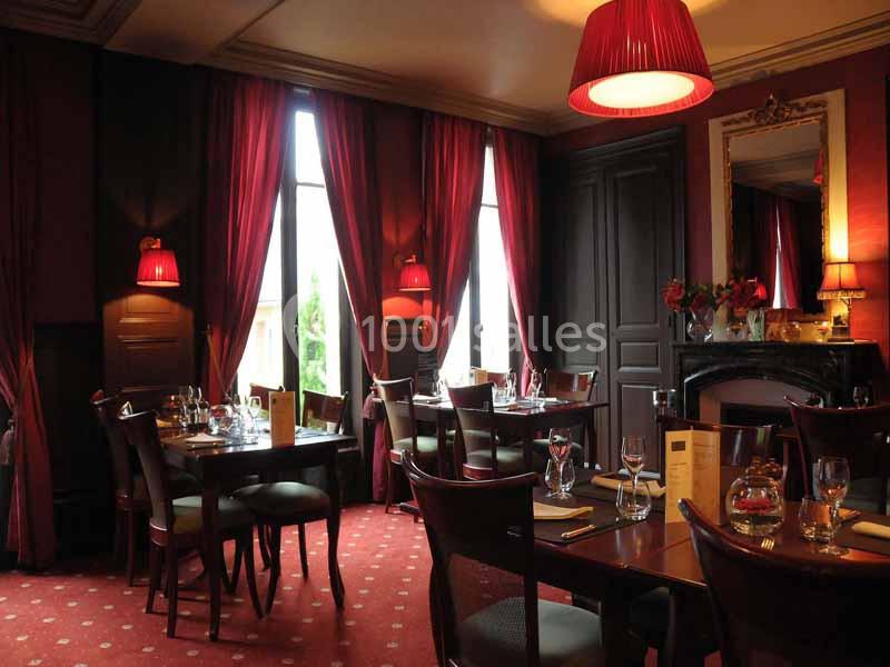 Salle de restaurant élégante avec tables dressées, rideaux rouges, éclairage tamisé et cheminée décorative.