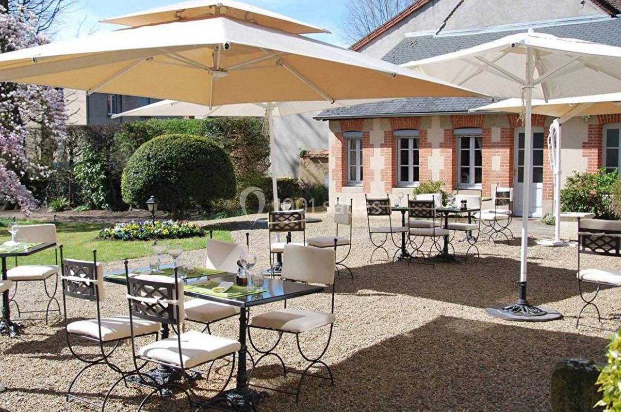 Terrasse extérieure avec tables et chaises en métal, parasols beige, jardin fleuri et bâtiment en arrière-plan.