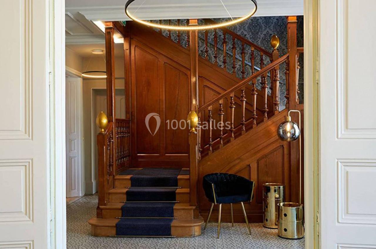 Escalier en bois avec rampe sculptée, tapis bleu, fauteuil noir et éléments décoratifs dorés dans un intérieur élégant.