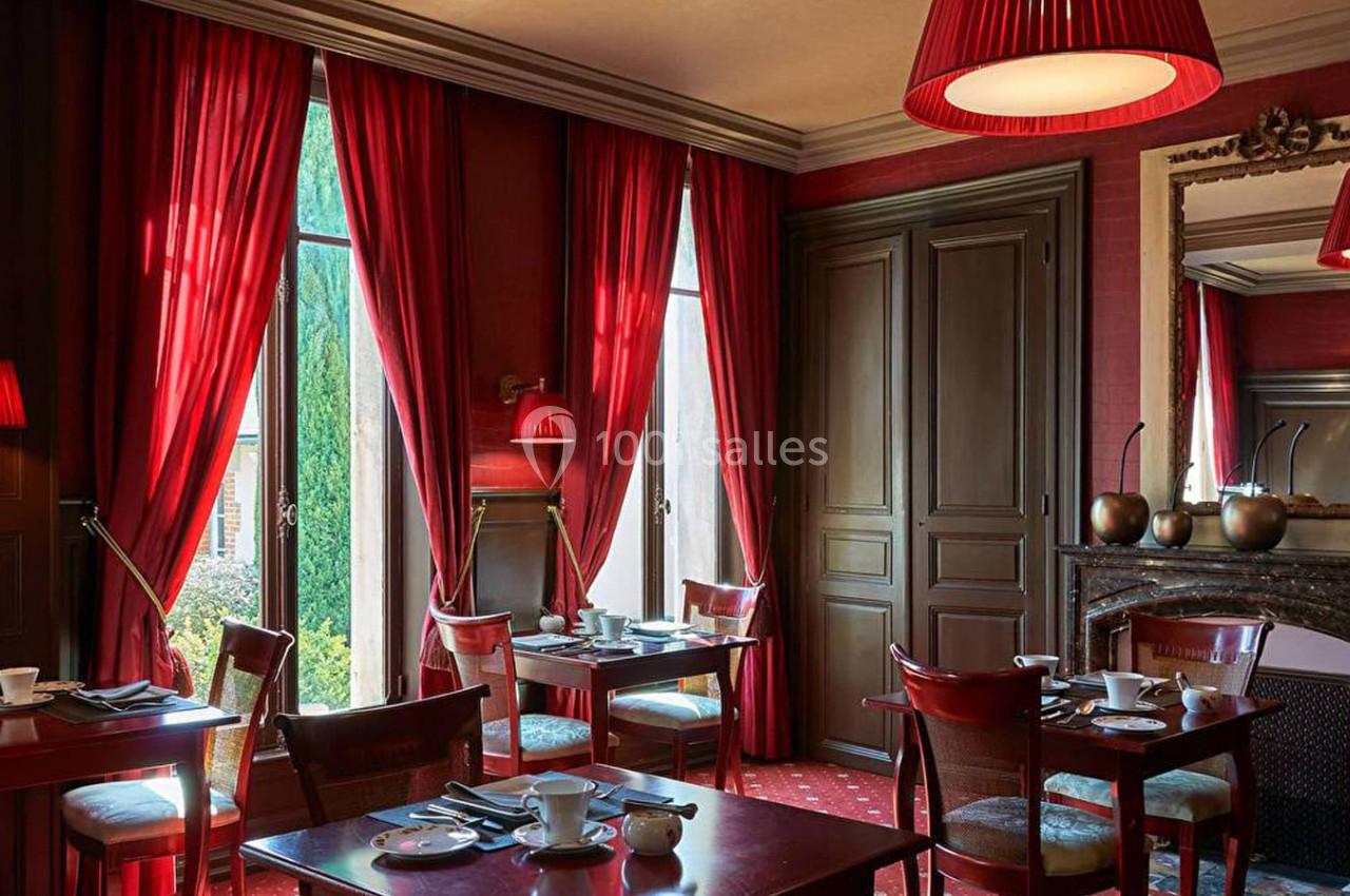 Salle à manger élégante avec rideaux rouges, tables dressées, cheminée et lumière naturelle entrant par de grandes fenêtres.