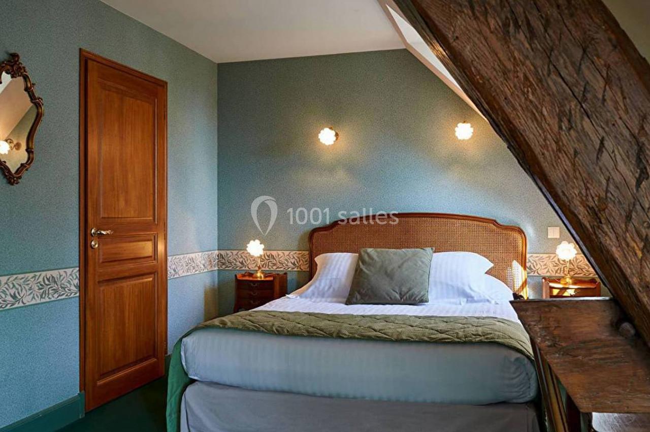 Chambre mansardée avec lit double, murs bleus, poutre en bois apparente et éclairage doux.