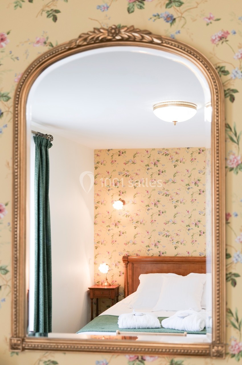 Reflet d'une chambre avec un lit double, des murs à motif floral et une lumière douce, vu dans un miroir doré.
