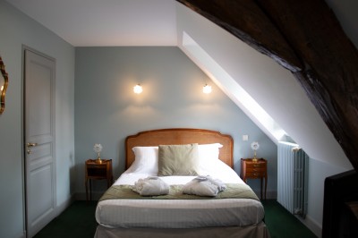 Chambre avec un lit double, draps blancs, coussins verts, deux tables de chevet et poutres apparentes au plafond.