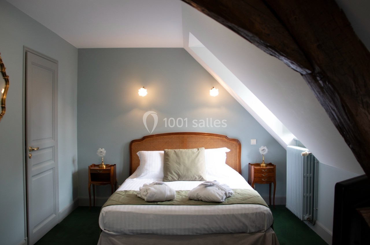 Chambre avec un lit double, draps blancs, coussins verts, deux tables de chevet et poutres apparentes au plafond.