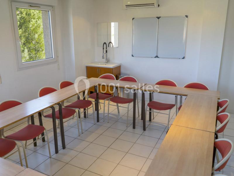 Salle de réunion lumineuse avec tables disposées en U, chaises rouges, tableau blanc et évier au fond.