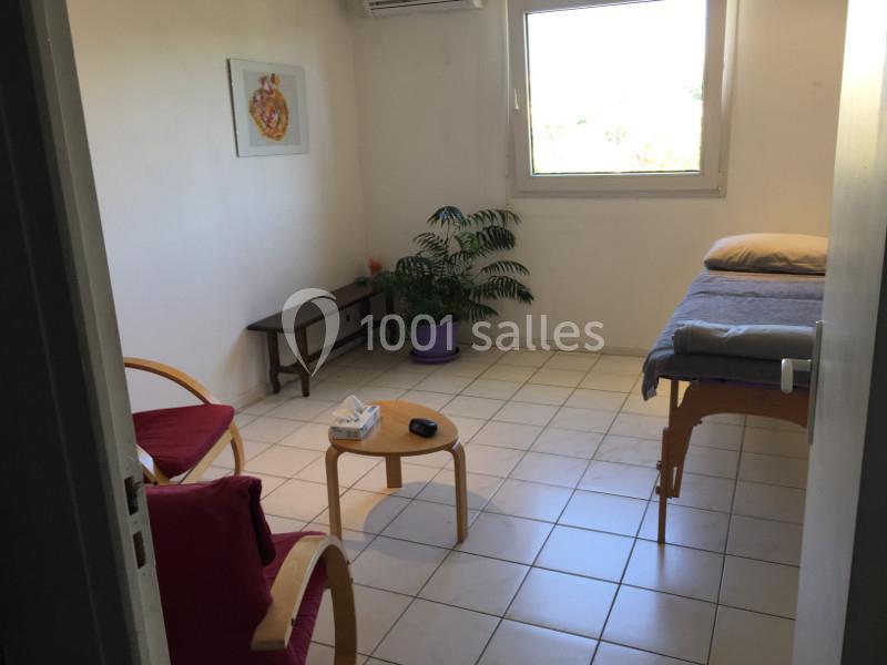 Salle lumineuse avec table de massage, chaises, petite table, plante en pot et fenêtre donnant sur l'extérieur.