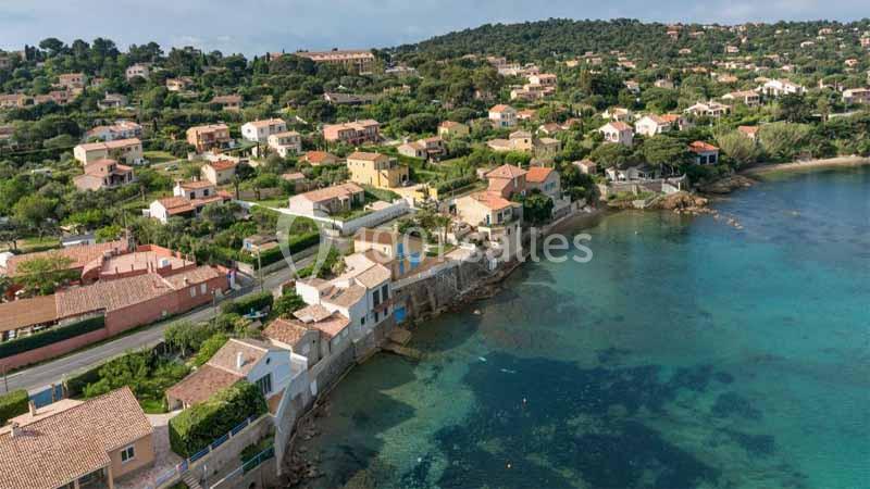 Vue aérienne d'un village côtier avec des maisons en bord de mer et une eau turquoise bordée de végétation.