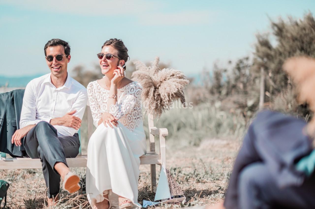 Un couple assis sur un banc en extérieur, souriant sous un ciel dégagé, entouré de végétation.