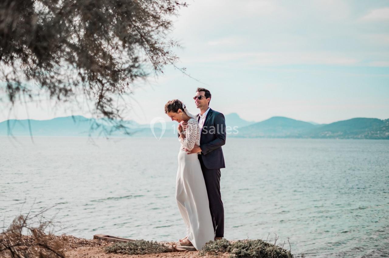 Un couple debout au bord d'un lac, l'homme en costume et la femme en robe blanche, entourés de nature.