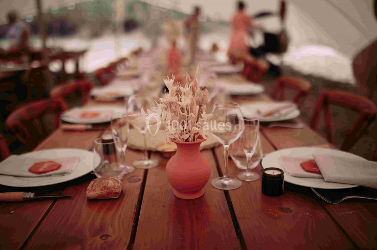 Table en bois dressée avec vaisselle, verres, serviettes et un vase central contenant des fleurs séchées.