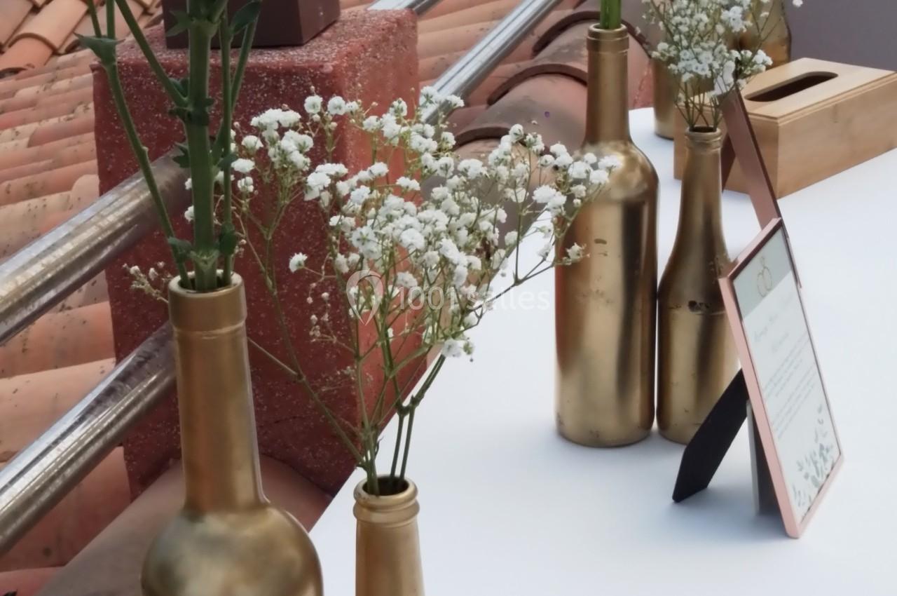 Bouteilles dorées recyclées servant de vases avec des fleurs blanches et vertes, disposées sur une table en extérieur.