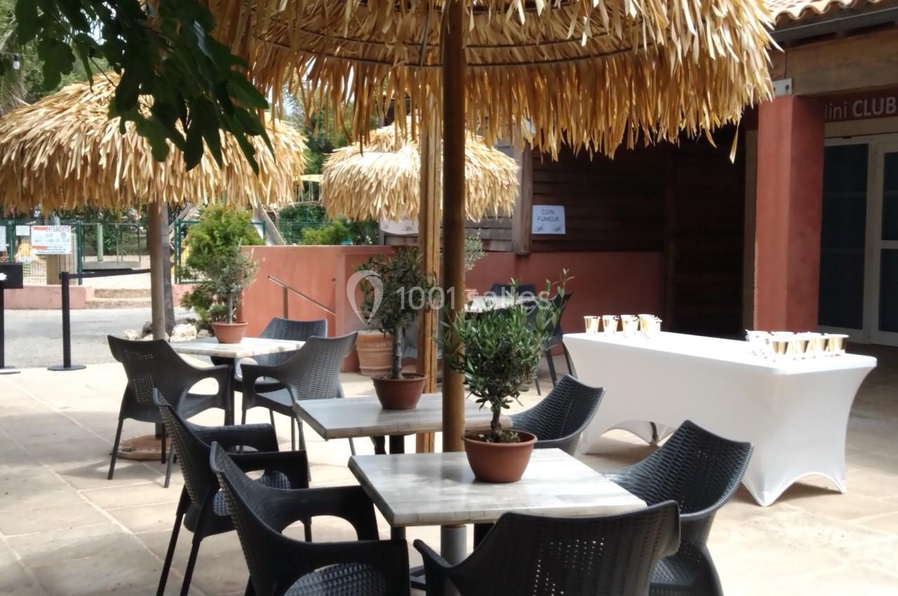 Terrasse avec tables rondes, chaises noires et parasols en paille, entourée de verdure et d'un bâtiment en arrière-plan.