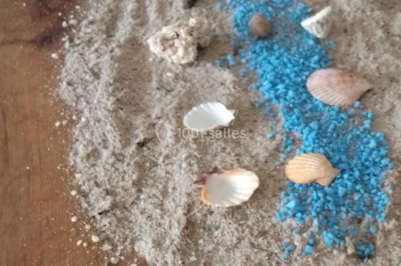 Décoration de table avec sable, coquillages, galets et un chemin de gravier bleu évoquant un cours d'eau.