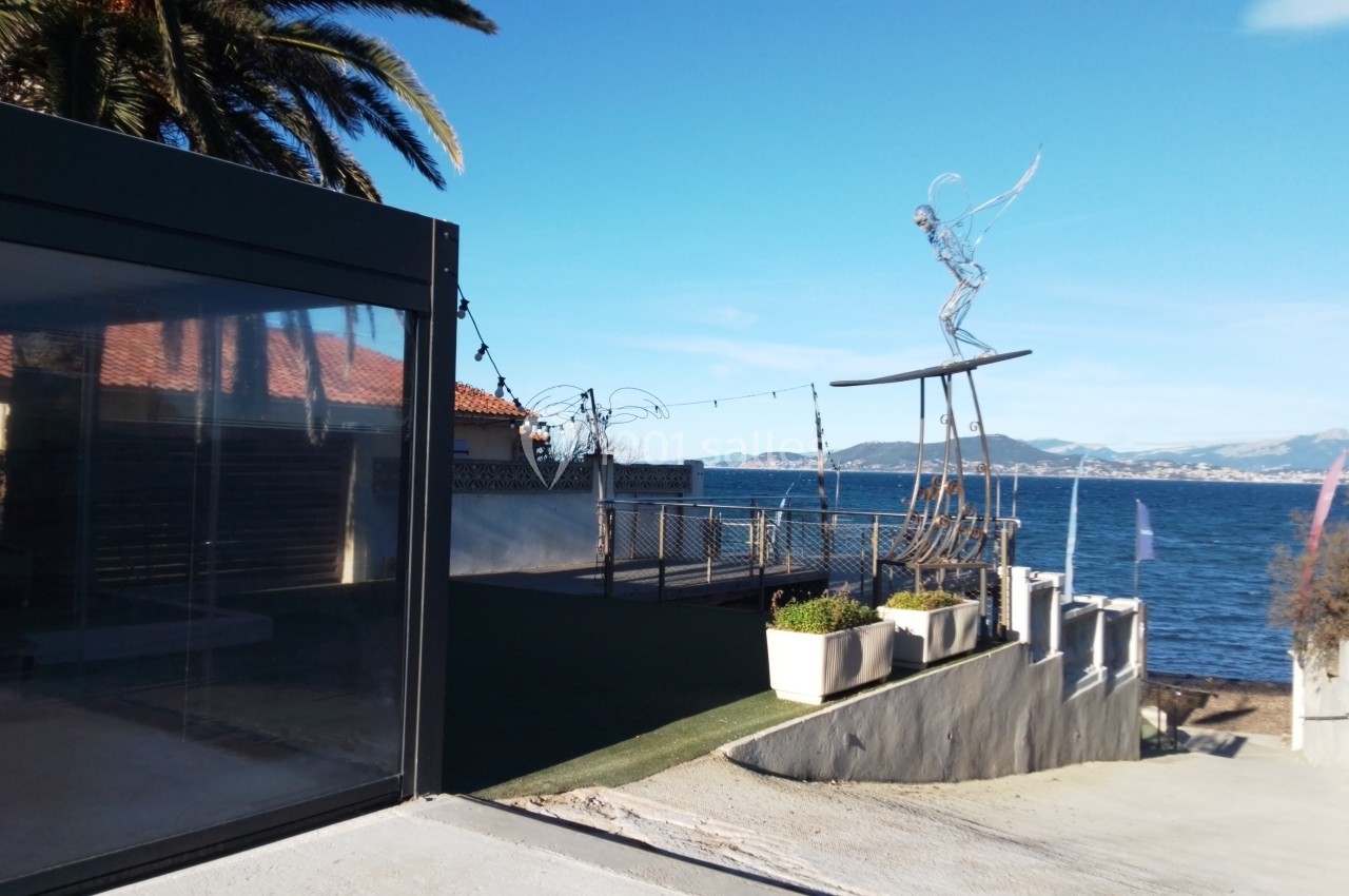 Vue d'une terrasse avec sculpture moderne, palmiers, mer et montagnes en arrière-plan sous un ciel bleu.