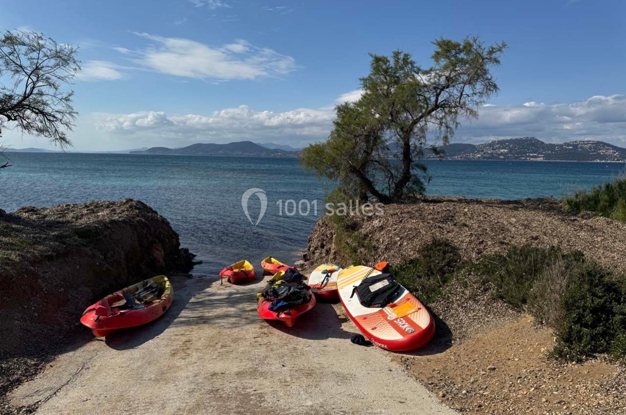 Des kayaks et une planche de paddle posés sur une rampe en béton menant à la mer, avec un paysage côtier en arrière-plan.