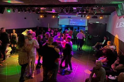 Des personnes dansent en couple sur une piste éclairée par des lumières colorées, tandis que d'autres sont assises autour.