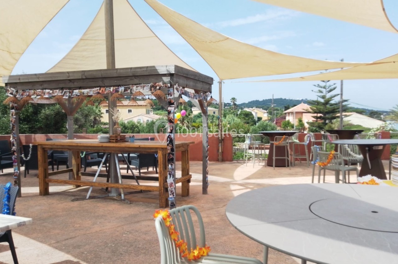 Terrasse extérieure avec tables, chaises et pergola ombragée, entourée de végétation et de maisons au loin.
