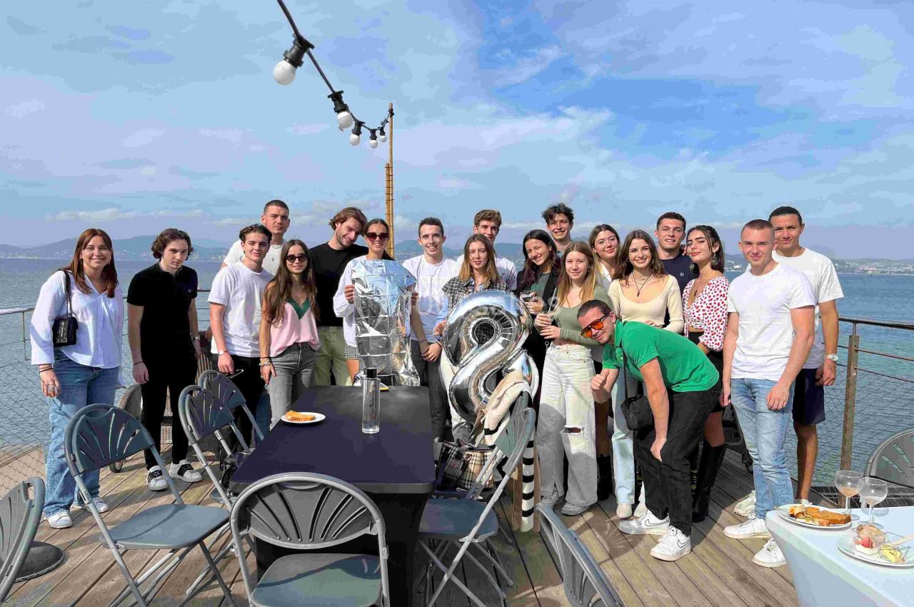 Un groupe de jeunes pose autour d'une table sur une terrasse en bord de mer, sous un ciel dégagé.
