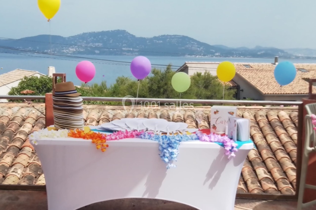Table décorée avec des colliers hawaïens et des ballons colorés en extérieur, vue sur des toits et un paysage côtier.