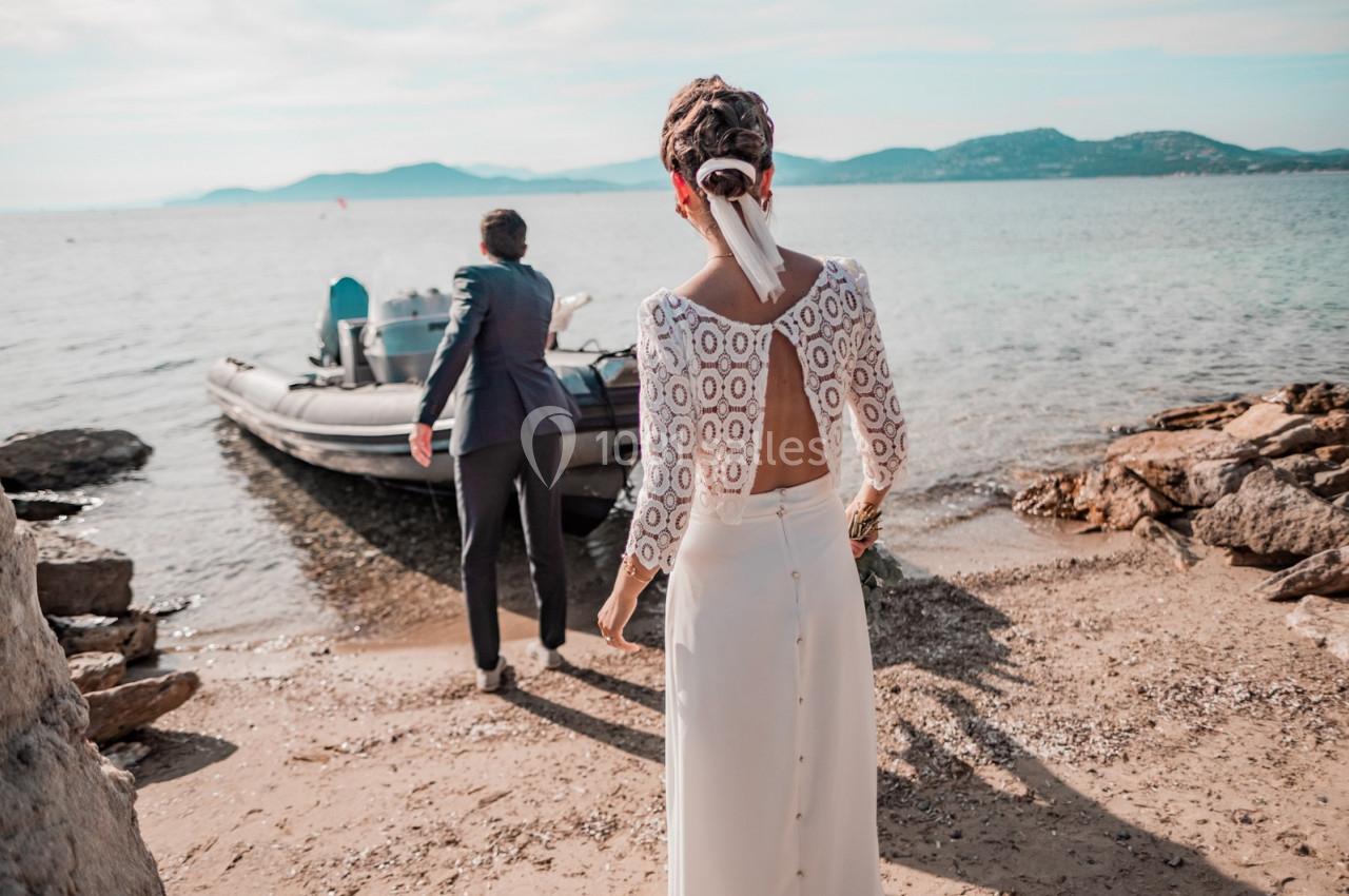 Une femme en robe de mariée marche vers un homme près d'un bateau pneumatique sur une plage.