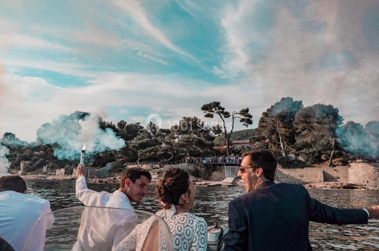 Des personnes sur un bateau célèbrent en allumant des fumigènes, avec une côte boisée en arrière-plan.