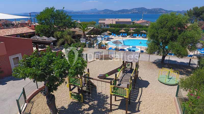 Aire de jeux pour enfants avec toboggan et structures en bois, près d'une piscine et vue sur la mer en arrière-plan.
